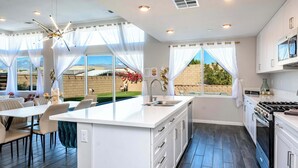 A modern kitchen with white cabinetry, a central island, and a dining area with chairs and a table. - @ Marbella Lane - Desert Dream • Pool/Spa & Games (Desert Hot Springs)