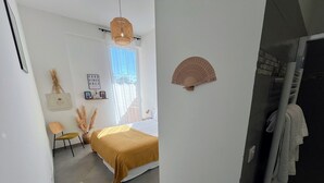A bedroom with a bed, a chair, a bedside table, a wall-mounted fan, and a window with curtains. - Beautiful seaside apartment with pool (Ajaccio)