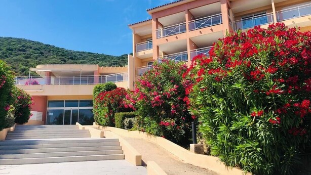 A multi-story building with a staircase leading to the entrance, surrounded by lush greenery and red flowers. - Beautiful seaside apartment with pool (Ajaccio)