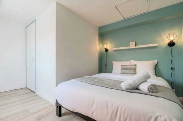 Un lit bien fait, avec du linge de lit blanc et un plaid gris, un mur d’accent de couleur turquoise et deux lampes de lecture fixées au mur.
