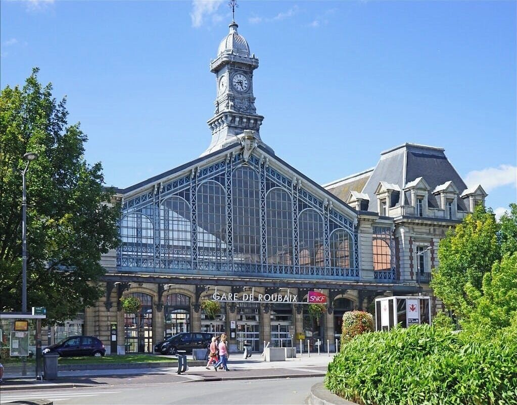 A historic building with a prominent clock tower, large glass windows, and a sign reading "GARE DE ROUBAIX".