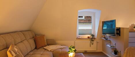 A cozy living room with a sofa, a coffee table with a plant, a TV mounted on the wall, and a window with blinds.