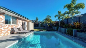 Une piscine avec des chaises longues, une maison en briques et un jardin avec différentes espèces de plantes.