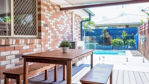 Un espace extérieur aménagé en patio, avec une table et des bancs en bois, un mur en briques et une vue sur une cour arrière avec une piscine et des plantes.