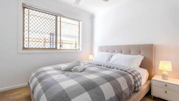 Een slaapkamer met een bed, nachtkastje, lamp, plafondventilator en een raam met uitzicht op gebouwen.