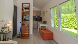 A compact room with a bed, a desk with a lamp, a bookshelf, a small table with a plant, and a window with a view of greenery.