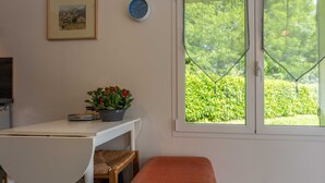 A room with a white table, a bench, a clock, a framed picture, and a window with a view of greenery.