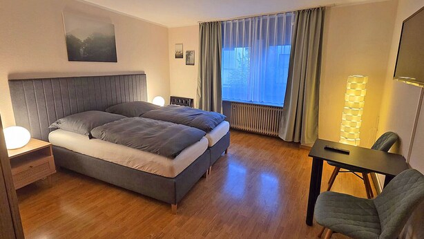 Una camera d'hotel con un letto grande, una scrivania con una sedia, una lampada da terra e una finestra con tende.