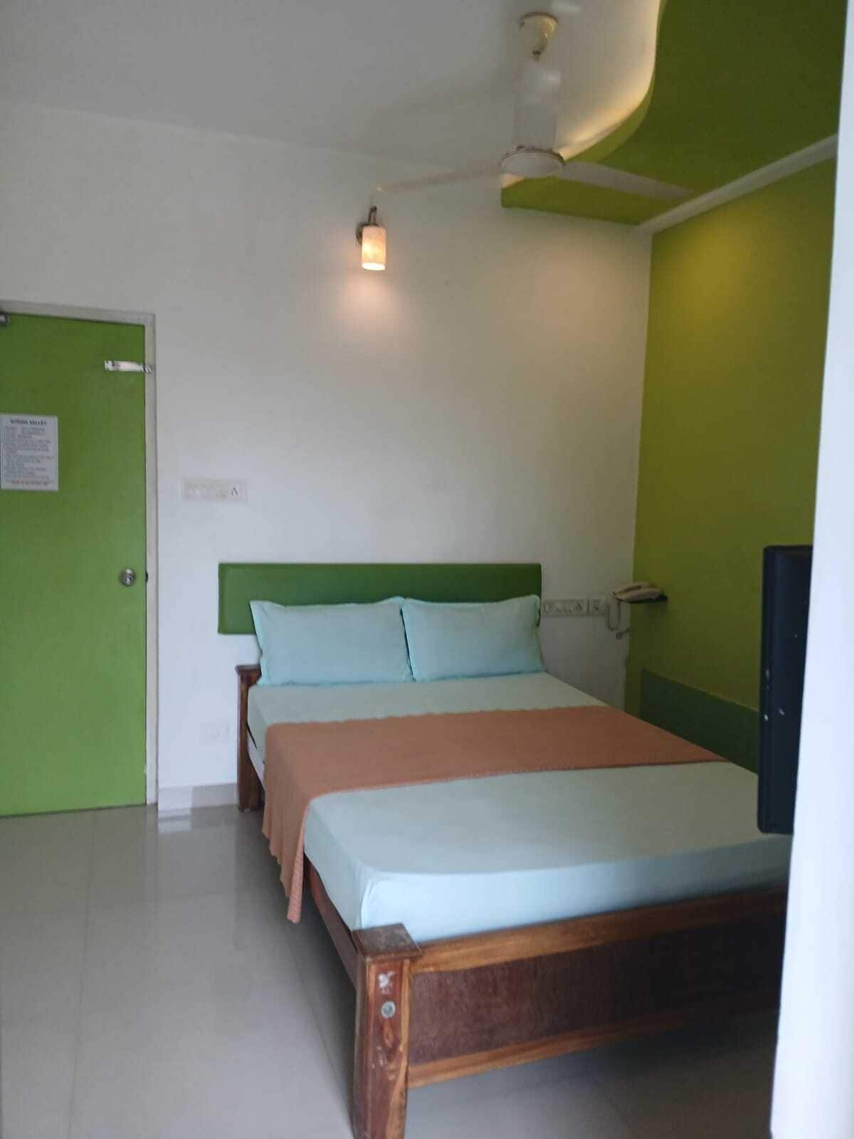 A single-bed room with a green door, a bedside table, a nightstand, and a wall-mounted lamp.