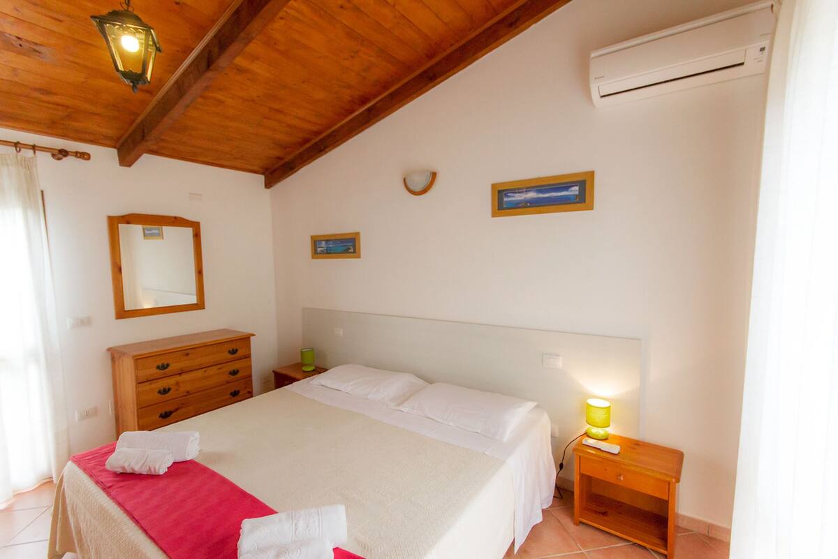 A bedroom with a wooden ceiling, a bed with white linens, a nightstand with a lamp, and a wall-mounted air conditioner.