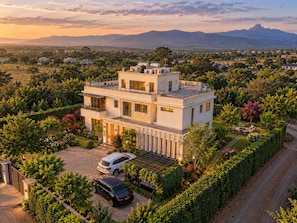 A modern two-story house with a flat roof, surrounded by lush greenery and a clear view of distant mountains.