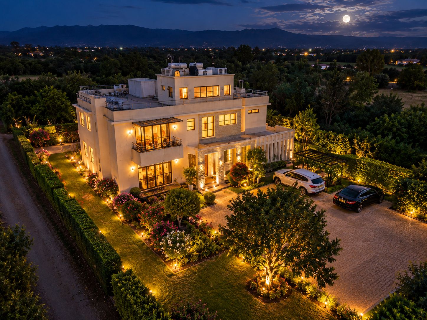 A modern two-story house with a well-lit driveway, surrounded by landscaped gardens and a clear view of the mountains in the background.