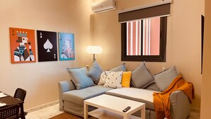 A living room with a sofa, a coffee table, and a lamp. There is a window with blinds and a wall-mounted air conditioner. - Relax family Élysée (Ouarzazate)