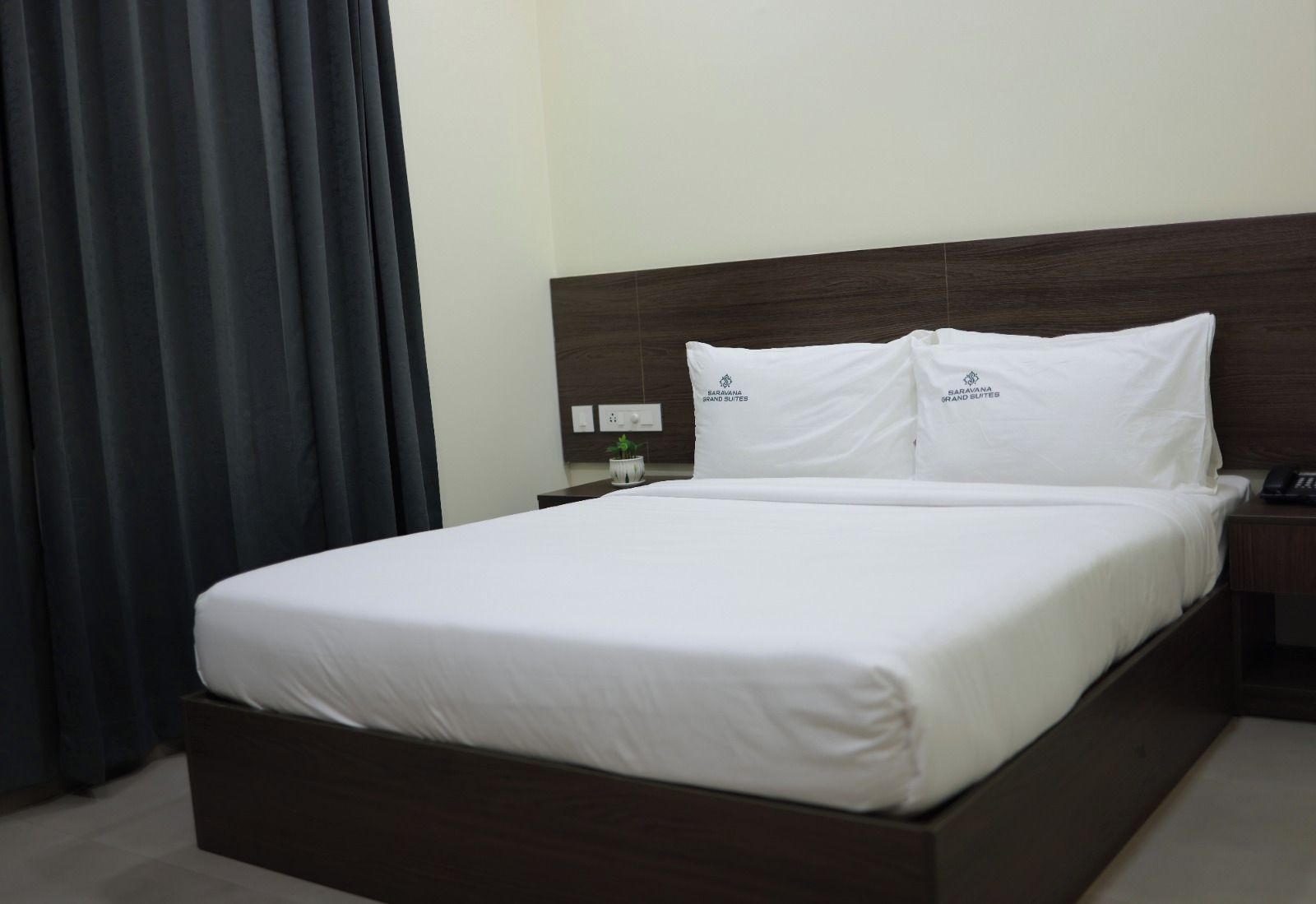 A hotel room with a large bed, white bedding, a dark wooden headboard, a nightstand with a phone, and a small potted plant.