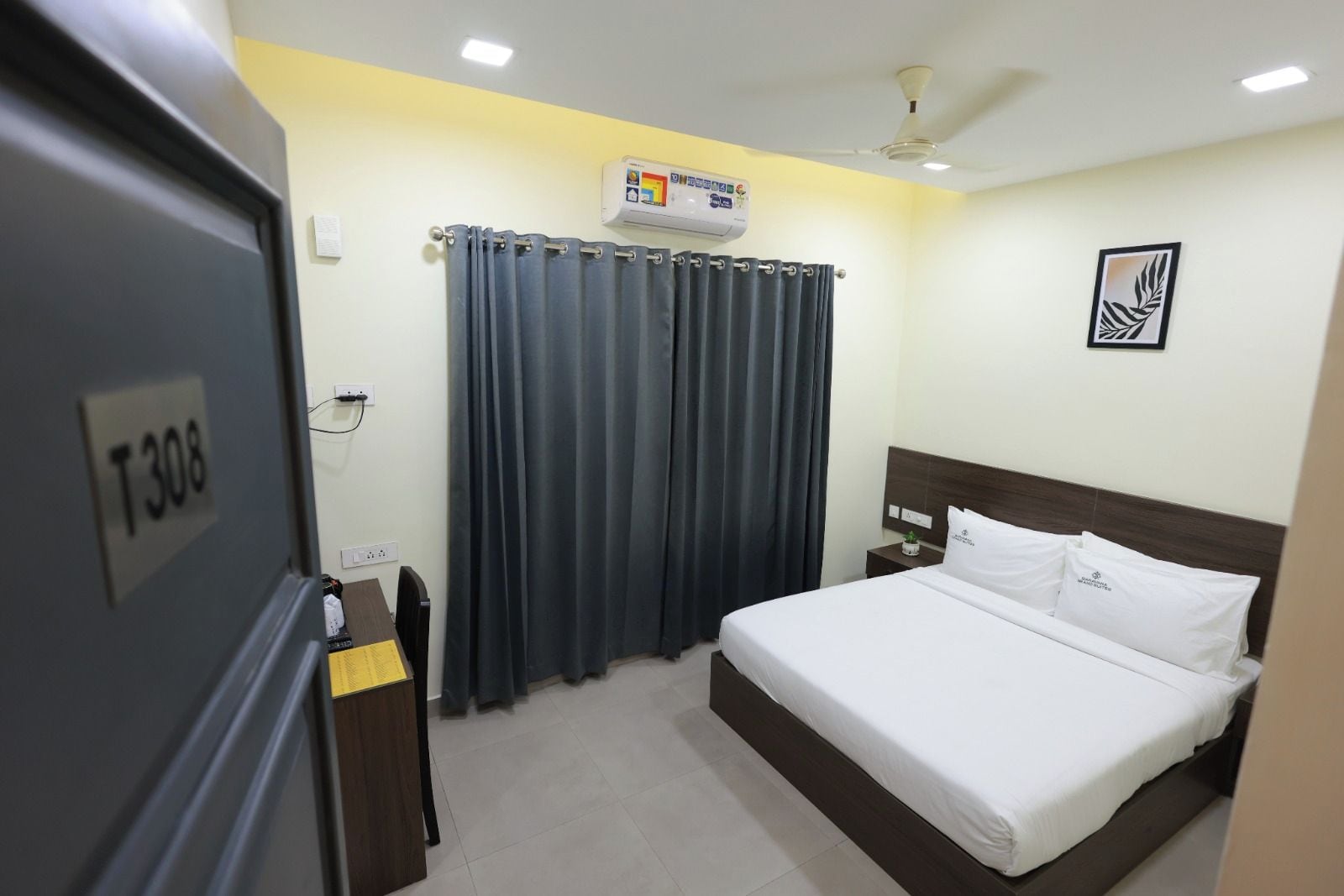 A hotel room with a bed, curtains, an air conditioning unit, a ceiling fan, and a wall-mounted artwork.