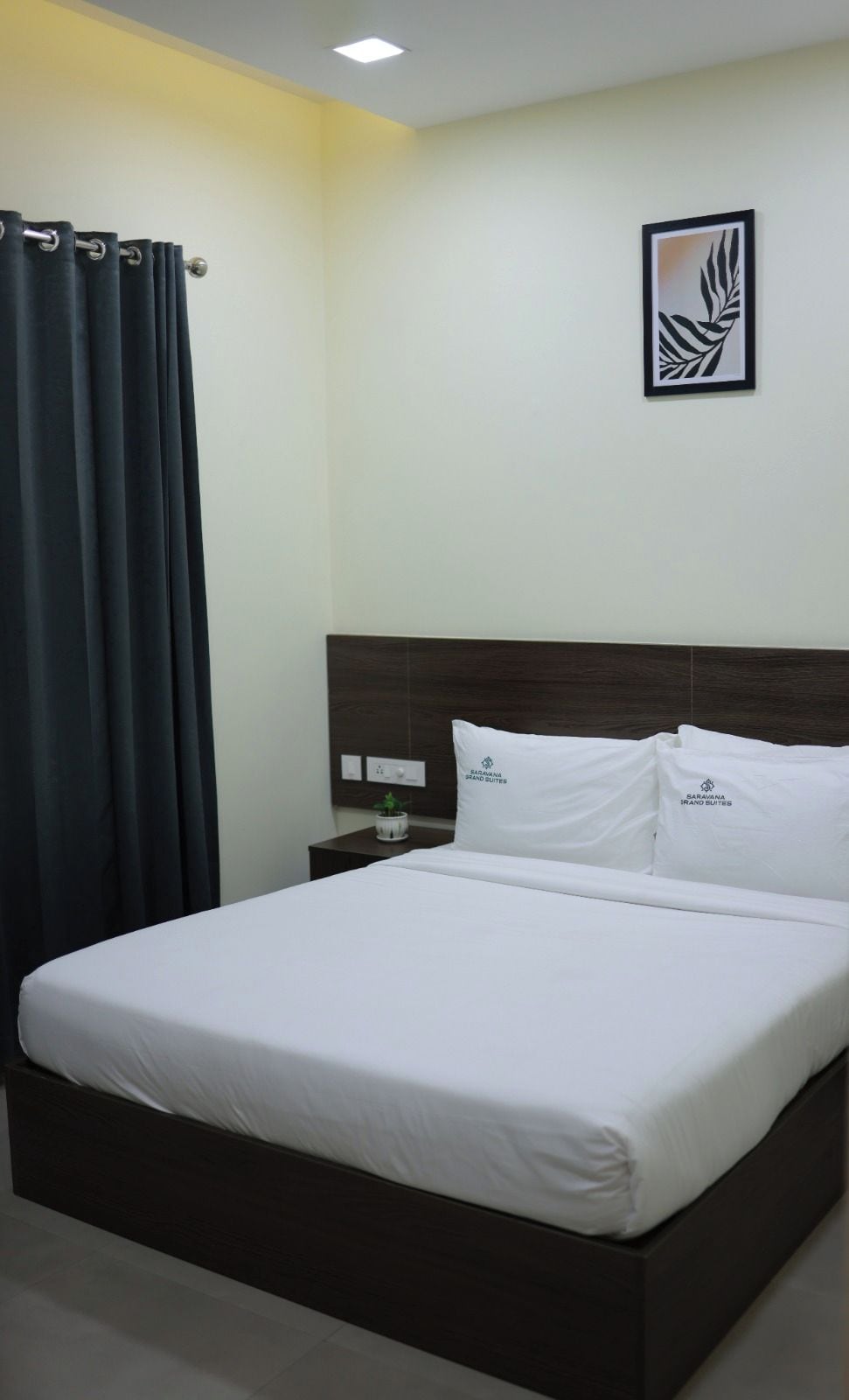 Ein Hotelzimmer mit einem Bett, dunklen Vorhängen, einem Nachttisch mit Pflanze und einem gerahmten Kunstwerk an der Wand.