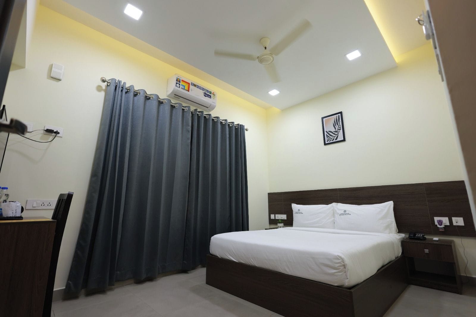 Une chambre d’hôtel comprenant un lit, une table de chevet, une commode, un ventilateur de plafond et un climatiseur fixé au mur.