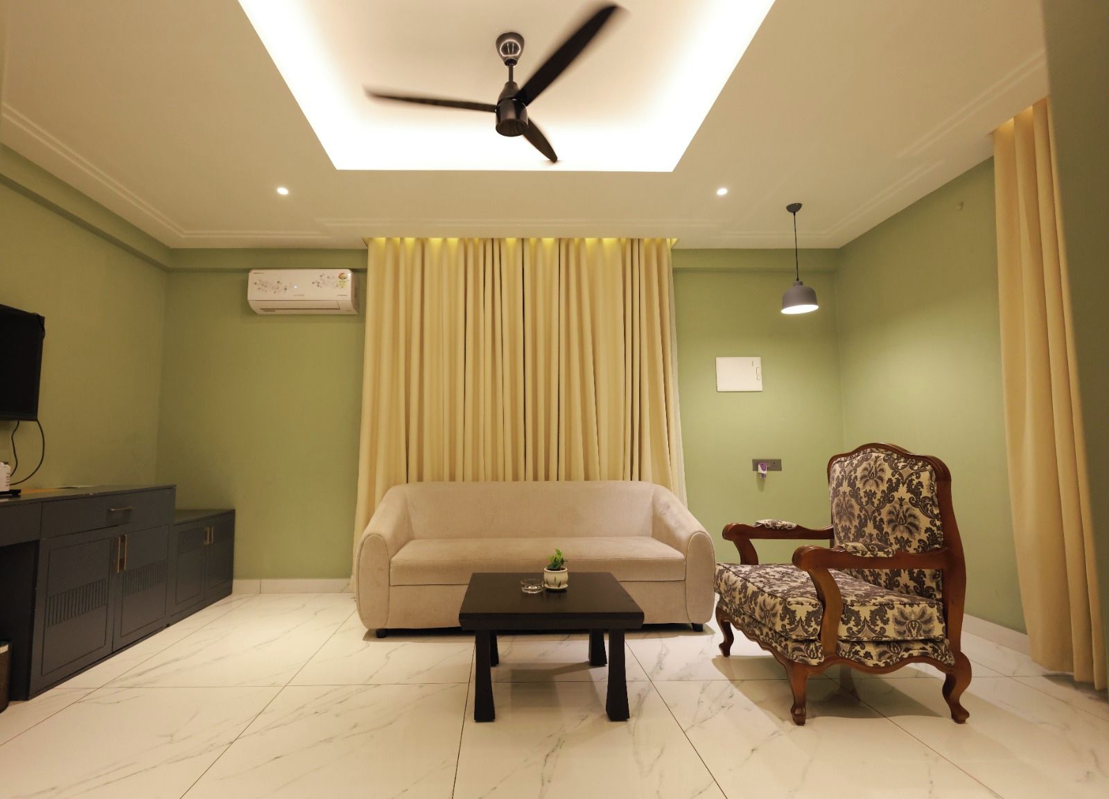 A living room with a sofa, a patterned armchair, a coffee table, a ceiling fan, and a television on a cabinet.
