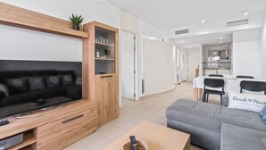 Un salon moderne doté d’un téléviseur à écran plat, d’un meuble de rangement en bois, d’un canapé gris et d’un coin repas avec une table et des chaises.