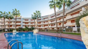 Une piscine avec des échelles en métal, entourée de palmiers et bordée d’un immeuble à plusieurs étages doté de balcons.