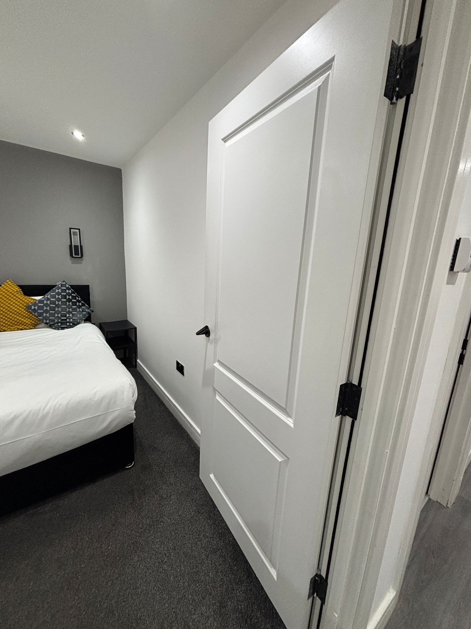 A bedroom with a bed, a nightstand, a bedside table, a door, and a wall-mounted light fixture.