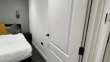 A bedroom with a bed, a nightstand, a bedside table, a door, and a wall-mounted light fixture.