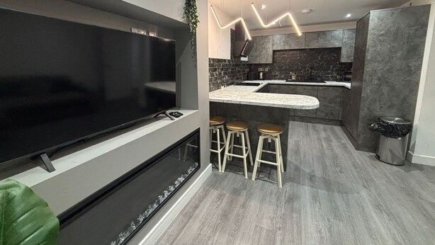 A modern kitchen with a large flat-screen TV, bar stools, and a marble countertop. - Aurora Apartments (Peterborough)