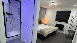 A modern bedroom with a built-in shower, a bed, a bedside table, and a window with blinds. - Aurora Apartments (Peterborough)