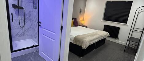 A modern bedroom with a built-in shower, a bed, a bedside table, and a window with blinds.