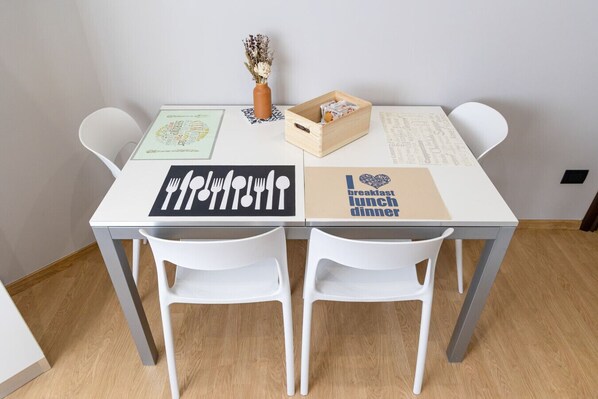 Una mesa blanca con dos sillas blancas, un cuadro con una cita, una caja de madera y un jarrón con flores secas.