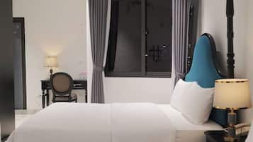 A hotel room with a large bed, a desk with a chair, a window with curtains, and a bedside table with a lamp.