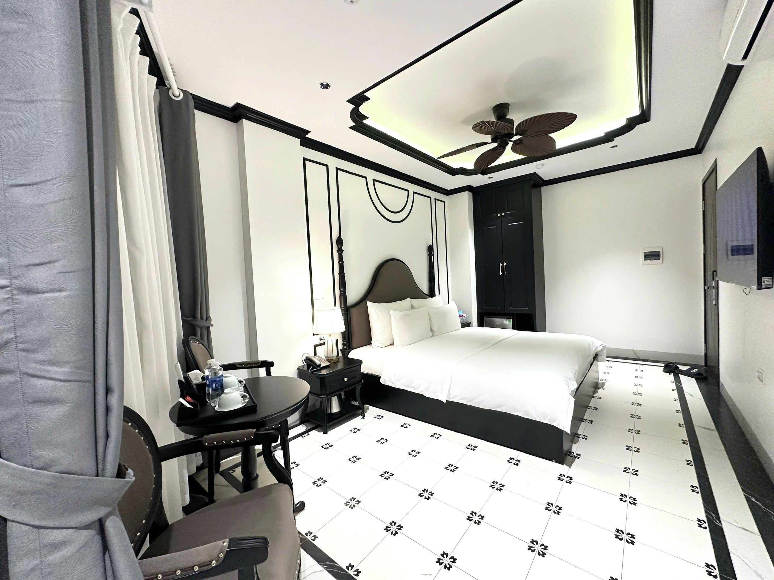 A hotel room with a bed, a desk with a chair, a desk lamp, a telephone, a water bottle, a cup, and a ceiling fan.