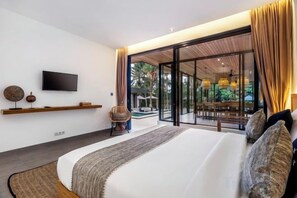 A modern bedroom with a large bed, a flat-screen TV, a dining area, and a view of the outdoors. - Villa Maya Bay by BaliSuperHost (Bali)