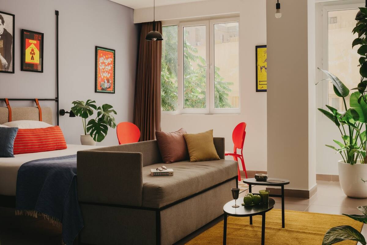 Ein modernes Wohnzimmer mit einer Couch, einem kleinen Tisch und einem Bett. Pflanzen, ein Fenster mit Vorhängen und gerahmte Bilder schmücken die Wand.
