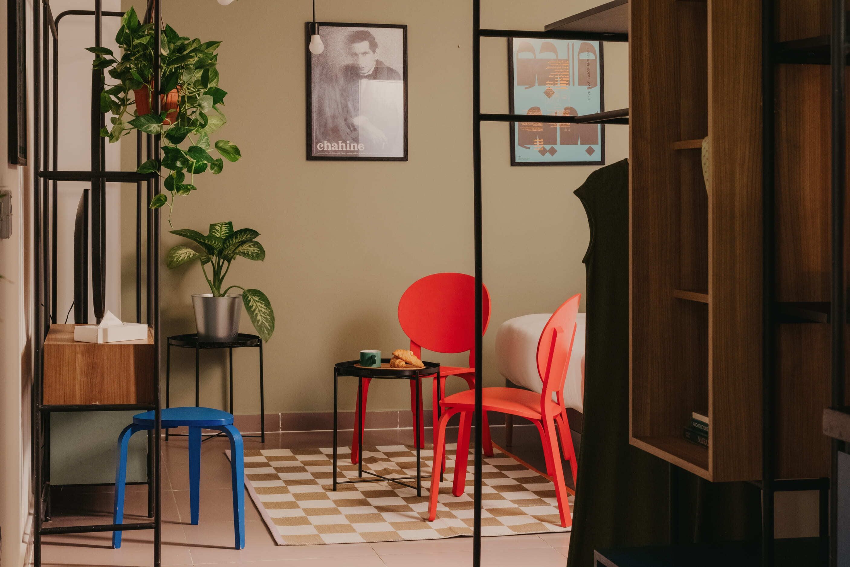 A room with a checkered floor, a blue stool, a red chair, a small table with a cup and a plate, and a potted plant.