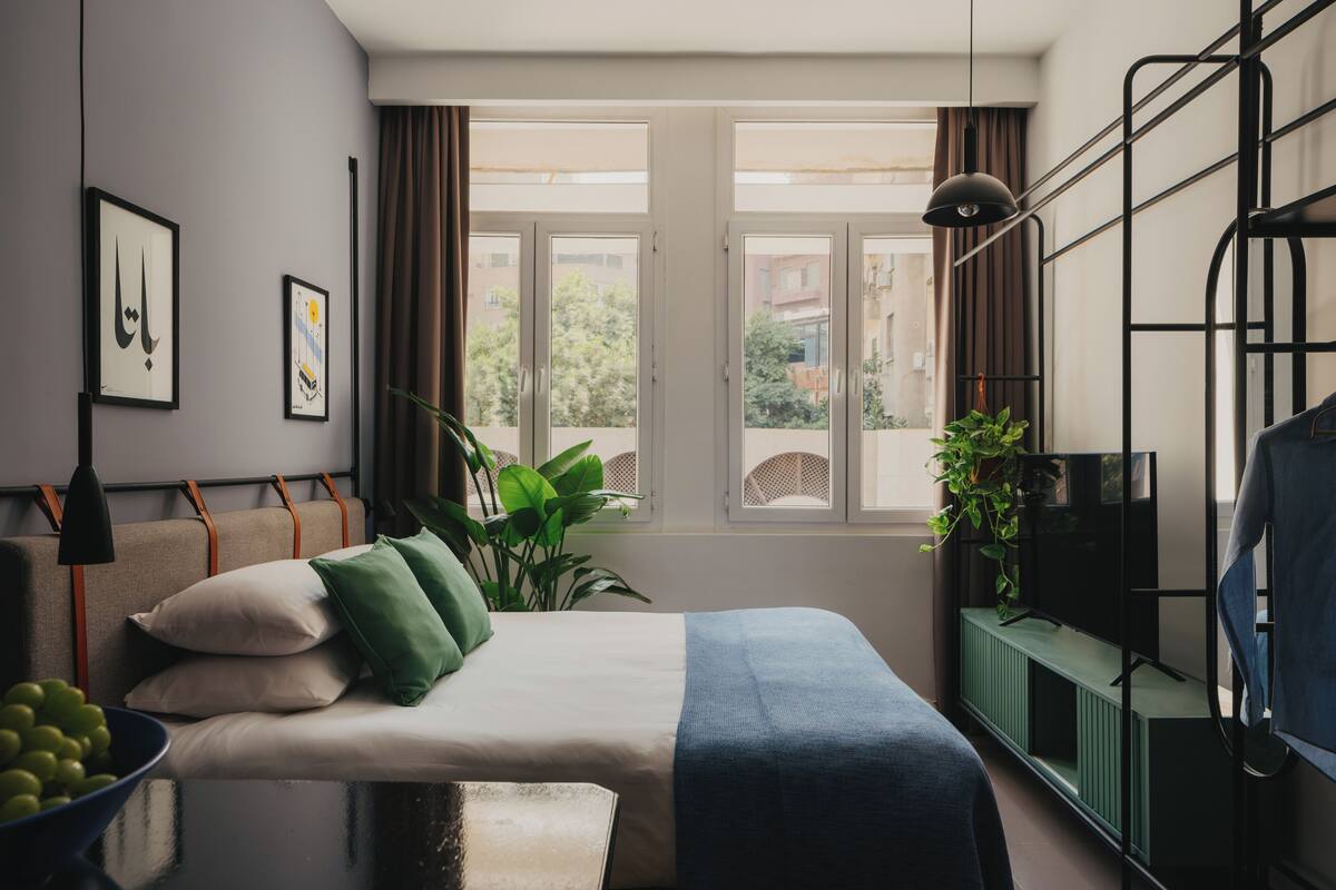 Ein modernes Schlafzimmer mit einem Bett, einem Fernseher auf einem Schrank, einem Fenster mit Vorhängen und einer Pflanze.