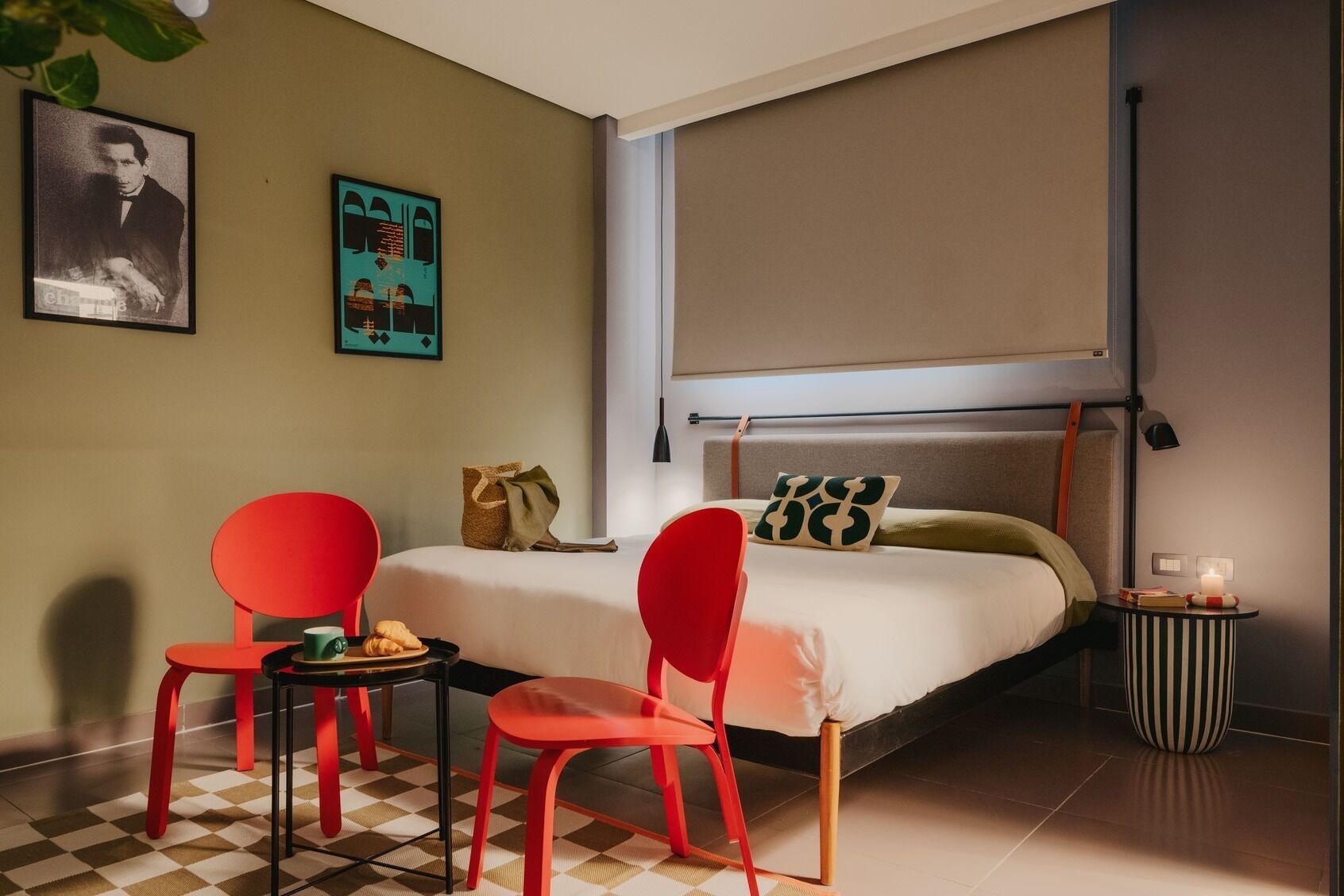 A hotel room with a bed, two red chairs, a small table with a croissant and a cup, a nightstand with a candle, and a window with a blind.