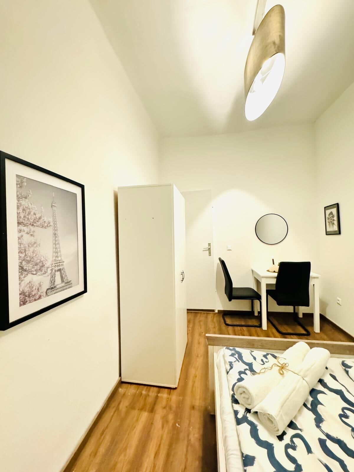 A hotel room with a bed, a desk, a chair, a framed picture of the Eiffel Tower, and a door.