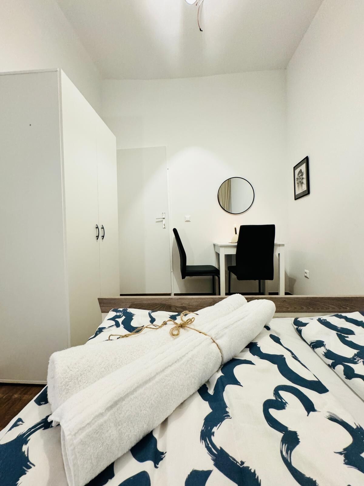A hotel room with a bed, a desk, a chair, a wardrobe, and a round mirror on the wall.