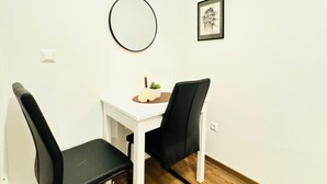 Una habitación pequeña y blanca, con piso de madera, una mesa blanca, dos sillas negras, un espejo redondo y un cuadro en la pared.