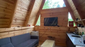 Tunnelmallinen ullakkohuone, jossa on puinen, kalteva katto, sohva, pieni pöytä, seinälle asennettu televisio sekä keittiöalue, jossa on pesuallas ja työtaso.