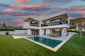 Una casa moderna de dos pisos con piscina, un patio con sillas y vistas al entorno verde.