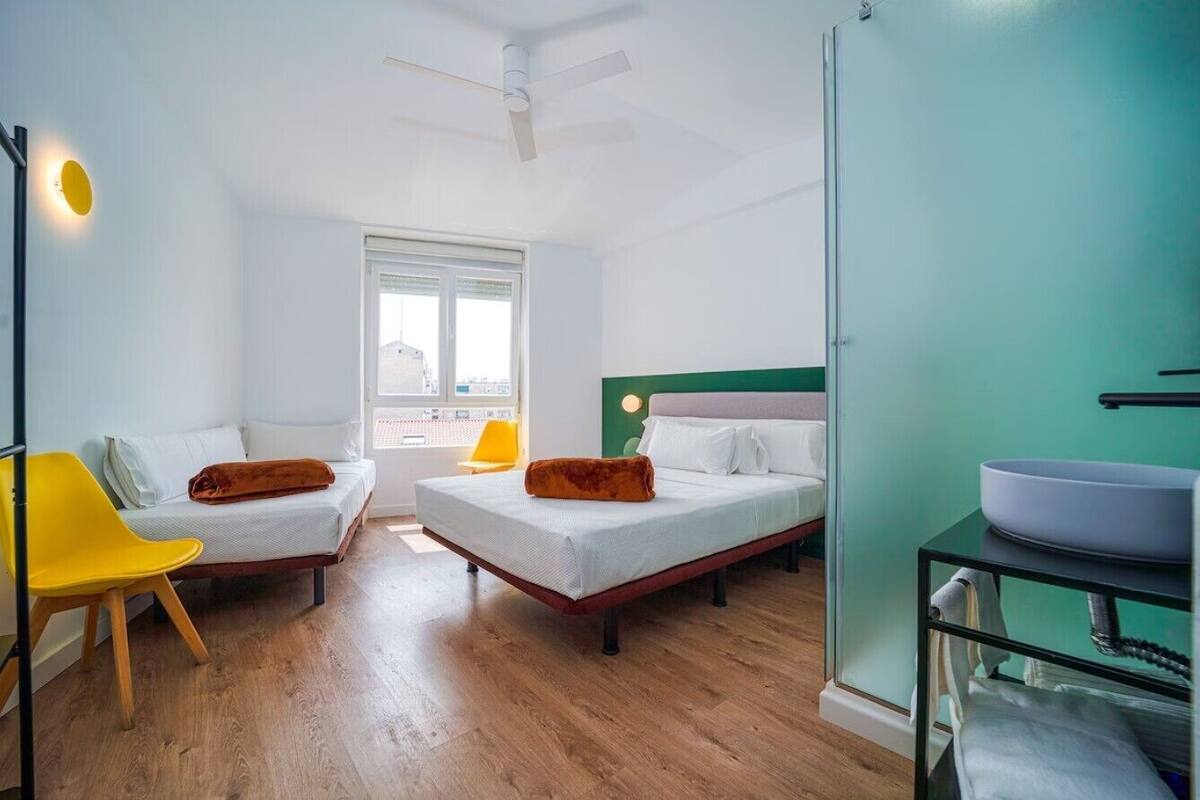 Een kamer met twee bedden, een gele stoel, een raam en een badkamer met een wastafel.