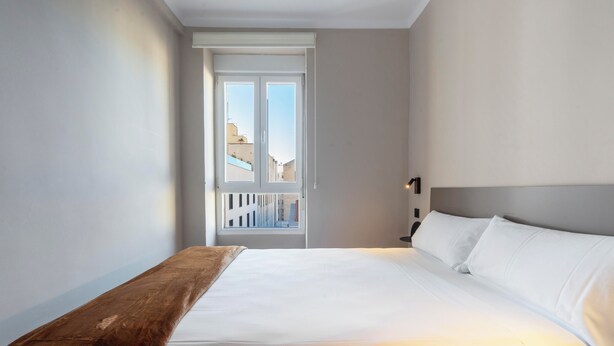Une chambre d’hôtel avec un grand lit, un banc en bois, une fenêtre donnant sur des immeubles et une table de chevet avec une lampe.