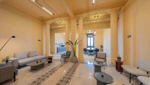 A spacious living area with a sofa, armchairs, a coffee table, and a dining table with chairs. There are potted plants and a large window providing natural light. - Brassbell Grey Studio CornicheAlexandria (Alexandria)