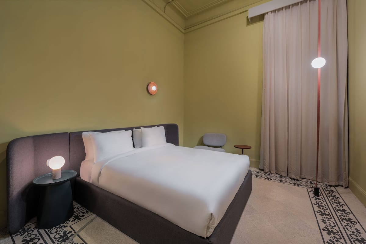 Een hotelkamer met een groot bed, een nachtkastje met lamp, een kleine stoel en een raam met gordijnen.
