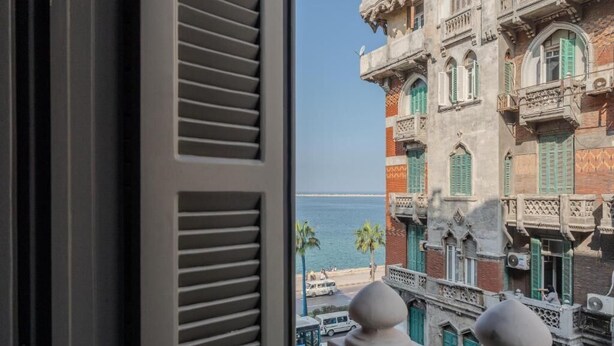 A view from a window with shutters, overlooking a coastal area with traditional architecture and palm trees. - Brassbell Grey Studio CornicheAlexandria (Alexandria)