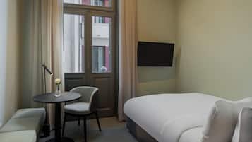 Quarto de hotel com uma cama, uma mesinha com uma cadeira, televisão na parede e janela com cortinas.
