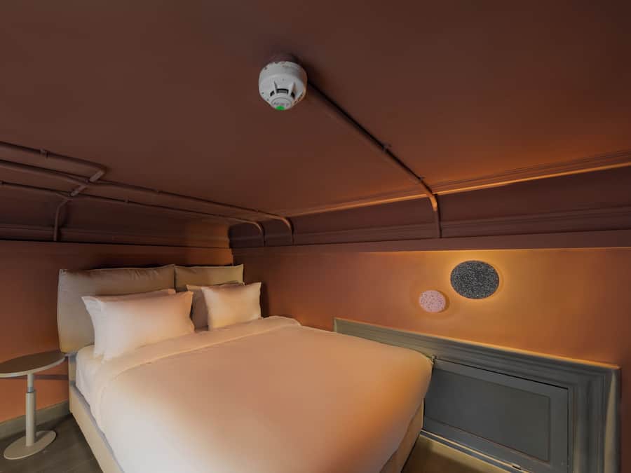 Een netjes opgemaakt bed met kussens, een nachtkastje, een wandlamp en een rookmelder.