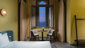A hotel room with a bed, two chairs, a small table with a plant, and a view of the sea through a window with open shutters. - Brassbell Grey Studio CornicheAlexandria (Alexandria)